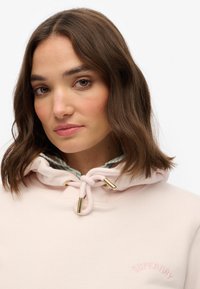 Sudadera con capucha de color rosa claro con herrajes dorados, cordón ajustable y forro a cuadros en azul. Presenta bordado "SUPERDRY" en el pecho.