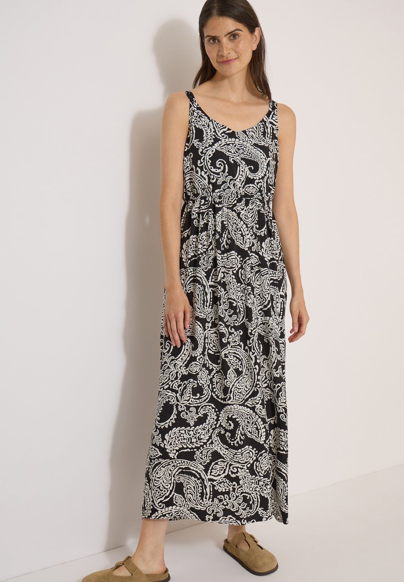 Femme debout contre un mur blanc, portant une robe longue noire sans manches à motif cachemire blanc et des sandales beiges à enfiler.