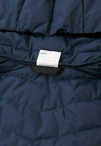 Wnętrze granatowej puchowej kurtki z pikowanym wzorem. Widoczna przyszyta etykieta z instrukcjami, zawierająca puste miejsce oraz wiadomość, aby przekazać ją dalej.