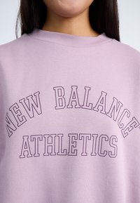 Sudadera lila con cuello redondo que presenta "NEW BALANCE" y "ATHLETICS" impresos en un tono púrpura más oscuro en un diseño arqueado.