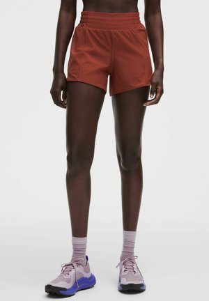 Jambes fines portant un short de sport couleur rouille, des chaussettes violet clair et des baskets assorties violet clair et bleu sur un fond clair uni.