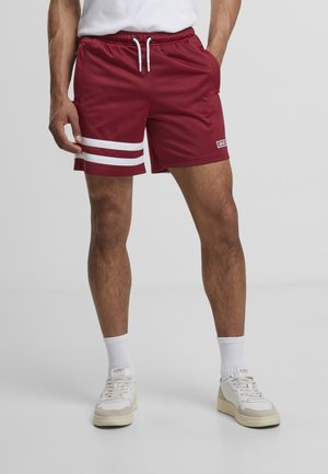 Hombre con pantalones cortos deportivos granate con rayas blancas en una pierna, calcetines blancos y zapatillas blancas, de pie sobre un fondo gris liso.