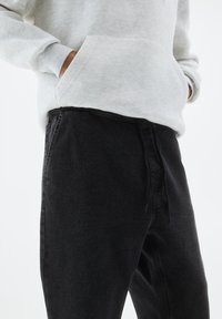 Felpa grigia chiara con tasca frontale, abbinata a pantaloni in denim neri con vestibilità dritta e cuciture visibili.