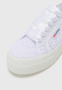 White lace sneaker with satin white laces, rubber sole, and textured detailing on the upper. Features a small red and blue logo.