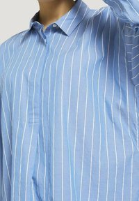 Ljusblå skjorta med knappar, vertikala vita ränder, krage och en enda bröstficka. Tyget verkar vara något strukturerat och andas.