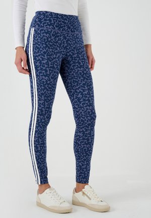 Mujer vestida con leggings de estampado animal azul con franjas laterales blancas y zapatillas blancas, de pie contra un fondo blanco liso.