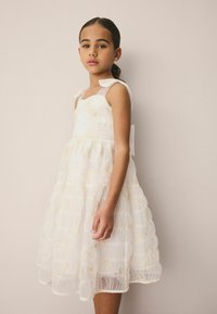 Ragazza giovane con un vestito bianco ricamato con motivi floreali, spalline trasparenti e dettagli a fiocco, in piedi contro uno sfondo beige uniforme.