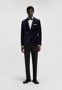 Giacca smoking in velluto blu navy con risvolti a punta, design monopetto, pantaloni neri, camicia bianca e papillon nero. Scarpe in vernice nere.