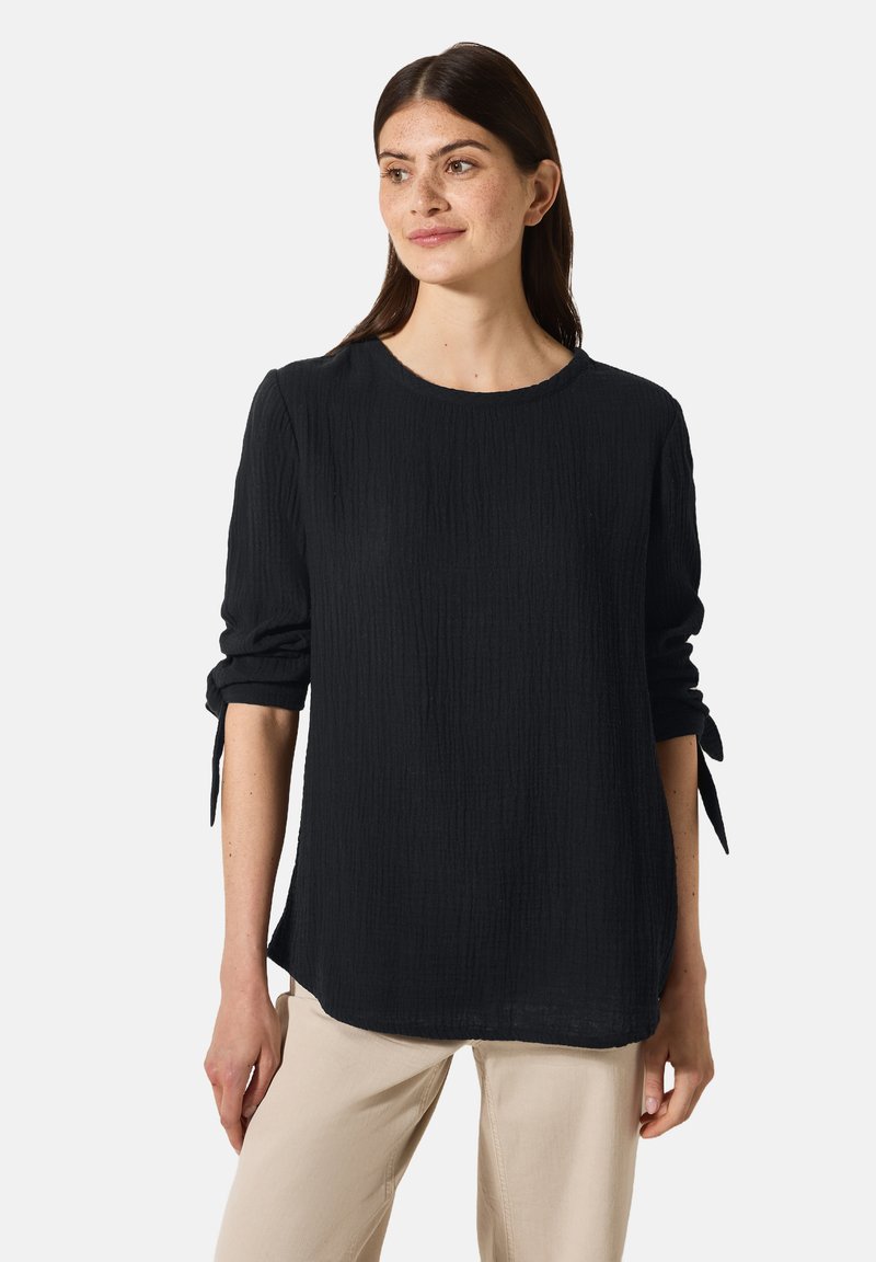 Chemise noire texturée à manches longues avec un col rond et des poignets noués, assortie à un pantalon beige clair. Design simple et décontracté.