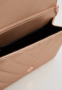 Rosaguld, vadderad, konstläderhandväska med tryckknappsstängning, fodrad inredning och synlig sömnad, som visar en stilren och modern design.