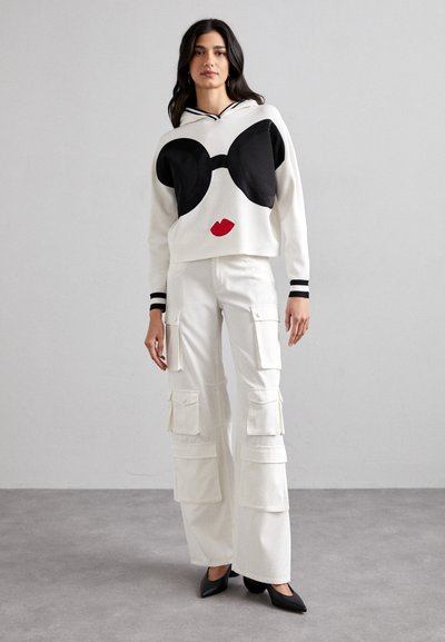 Pull avec design graphique noir et rouge évoquant des lunettes de soleil et des lèvres, associé à un pantalon cargo blanc comportant plusieurs poches.