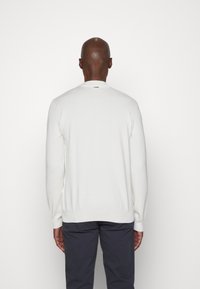 Pull en tricot blanc avec un col rond, des manches longues et un ourlet côtelé. Le dos affiche une étiquette logo discrète près de l'encolure.