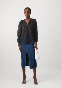 Blouse en dentelle noire à manches longues, décolleté en V et motif floral, associée à une jupe en denim longueur genoux avec une fente devant. Sac à main blanc.