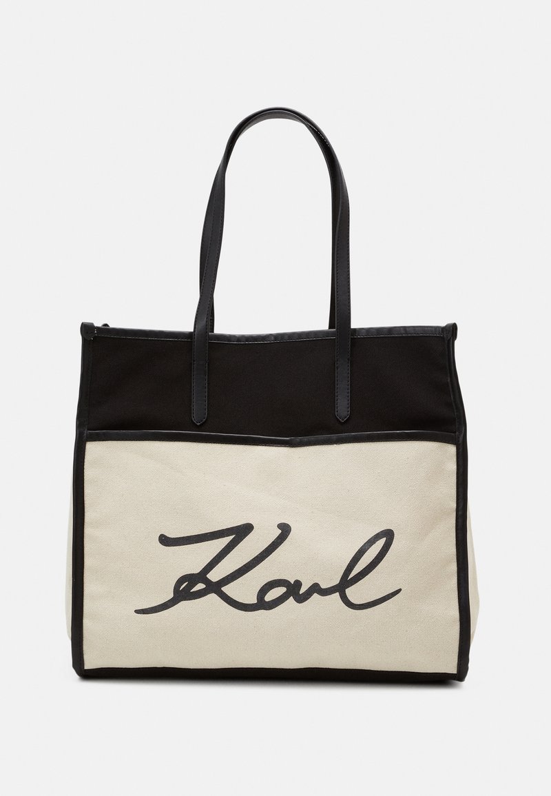 Sac fourre-tout en toile crème et noir. Il dispose de deux poignées noires, d'un panneau inférieur crème avec un logo en script noir, et d'un design structuré.