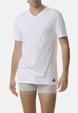 Male wearing a white V-neck T-shirt with a small Adidas logo and light gray fitted shorts against a plain background.
