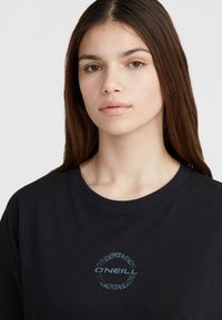 Sort bomulds-sweatshirt med ribbestrikket rund hals, der har et cirkulært blåt grafisk logo med tribal-accenter i midten.