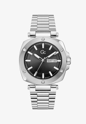 Strieborné kovové náramkové hodinky s čiernym ciferníkom, striebornými hodinovými indexmi, zobrazením dňa a dátumu s ukazovateľom SOB 14 a značkou „Gc“ pod pozíciou 12 hodín.