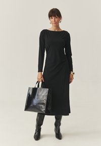 Rochie lungă neagră cu mâneci lungi, gât bateau, detalii drapate, asortată cu o geantă din piele neagră lucioasă și cizme negre până la genunchi.