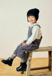 Salopette en velours côtelé gris à pois noirs, chemise blanche à manches longues, bonnet en tricot noir et bottes noires à la cheville avec des chaussettes grises, assise sur un tabouret en bois.
