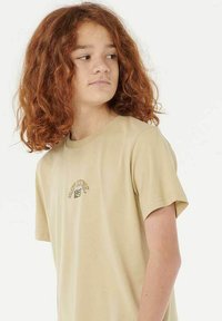 Camiseta de algodón color beige con mangas cortas, que presenta un pequeño diseño gráfico colorido en el pecho. Textura suave, ajuste relajado.