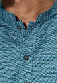 Blauw textuurshirt met zwarte knopen en een korte mandarin kraag op een persoon met een gemiddelde bruine huid.