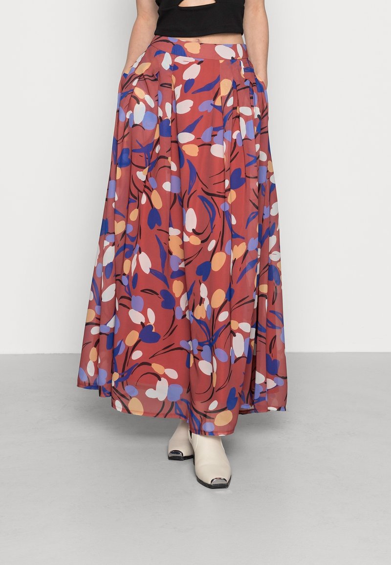 Maxihame punaisena, jossa on monivärinen kukkakuosi. Valmistettu kevyestä kankaasta, jossa on laskoksia ja taskuja. Päällä avonaiset kengät.