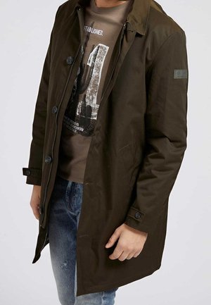 Homme portant un manteau boutonné marron foncé sur un t-shirt graphique marron et un jean bleu, main détendue sur le côté contre un fond clair uni.
