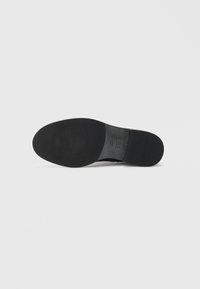 Semelle en caoutchouc noir avec une texture lisse et une forme arrondie. Présente des marques de la marque et un léger talon pour la stabilité.