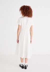 Rochie albă, până la genunchi, cu mâneci scurte și talie strânsă. Purtată cu pantofi negri și șosete albe până la gleznă, evidențiind un design simplu.