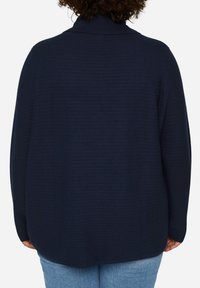 Maglione blu navy, con texture e colletto alto, caratterizzato da una vestibilità ampia e orlo arrotondato. La vista posteriore mostra la lunghezza completa e i dettagli delle maniche.