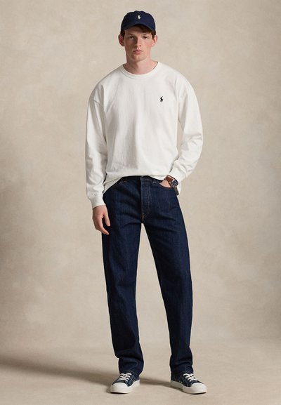 Νέος άνδρας που φοράει μπλε σκούφο, λευκό μακρυμάνικο πουκάμισο με λογότυπο, σκούρο μπλε τζιν και μπλε αθλητικά παπούτσια, στέκεται μπροστά σε μπεζ φόντο.