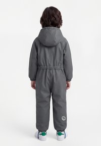 Kind draagt een donkergrijze jumpsuit met capuchon en elastische manchetten, met een rond logo nabij de linker enkel, staand met de rug naar de camera op een witte achtergrond.