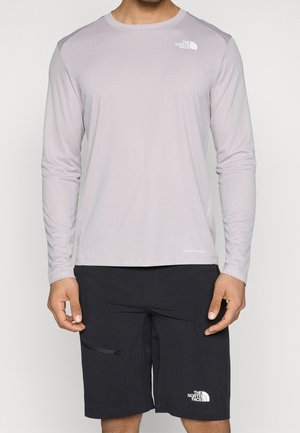 Mężczyzna w jasnoszarej, długorękawowej koszuli North Face oraz czarnych krótkich spodenkach North Face z zamykanym na zamek błyskawiczny bocznym kieszonką, stojący na jednolitym tle.