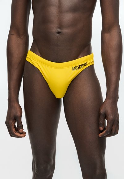 Άνδρας που φοράει φωτεινά κίτρινα μαγιό μπριφ με το λογότυπο «MOSCHINO» στο δεξί ισχίο, εμφανιζόμενος από το μέσο του κορμού μέχρι τους άνω μηρούς.