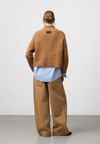 Pull court marron en matière douce, superposé sur une chemise bleu clair, associé à un pantalon large beige. Étiquette noire à l'arrière.