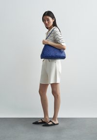 Kék, fonott válltáska hobo formával, állítható pánttal és sima textúrával. Csíkos felsővel és világos rövidnadrággal viselve, fekete lapos cipővel kiegészítve.
