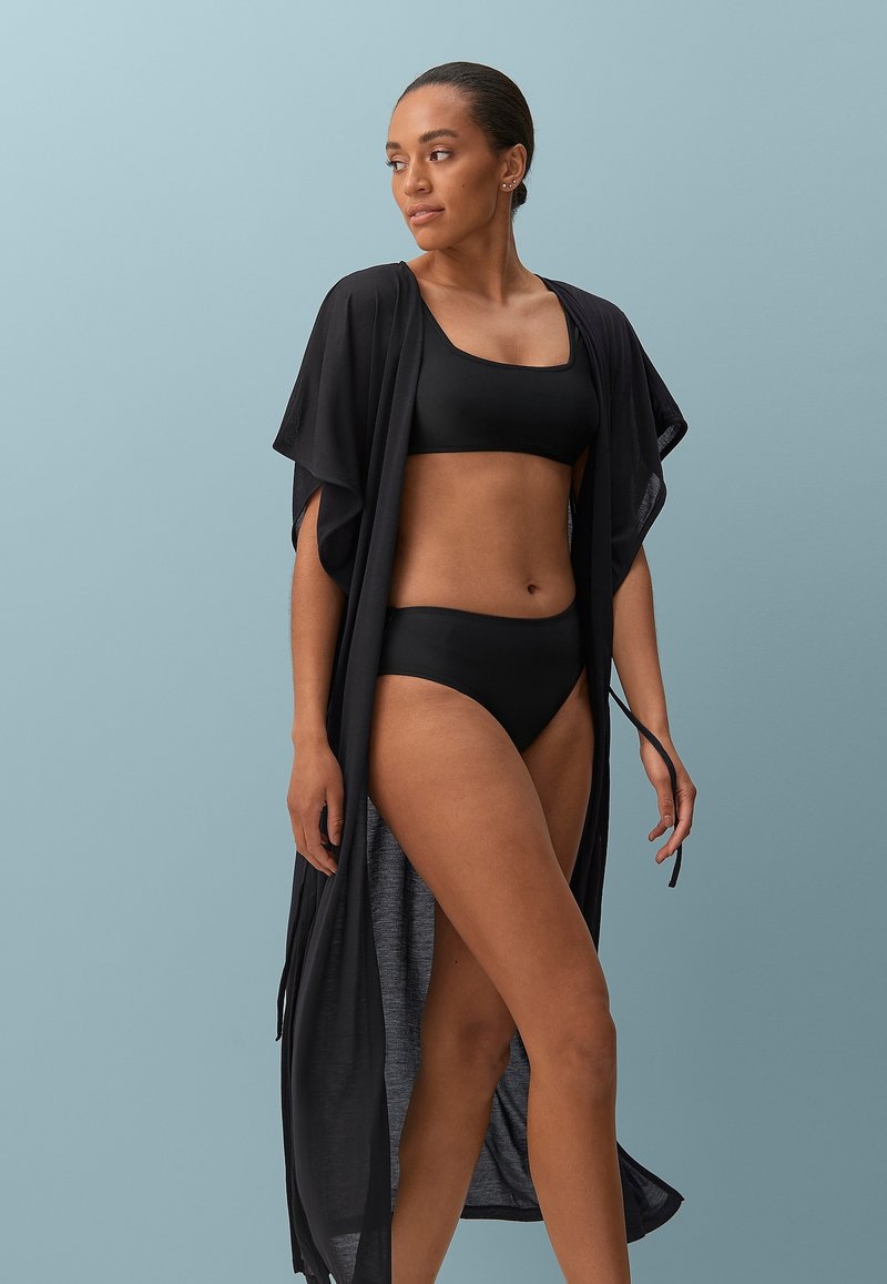 Zwart zwemsetje gelaagd met een lichtgewicht, doorzichtige zwarte kimono met brede mouwen en zijsplitten, tegen een lichtblauwe achtergrond.