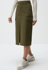 Olijfgroene midi-jurk gemaakt van katoenmix, met een uitsparing aan de voorkant en grote zijzakken, gecombineerd met witte sneakers.
