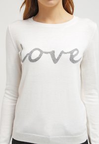 Witte lange mouwen sweater met het woord "liefde" in grijze cursief over de borst, gedragen door een persoon met lichtbruin haar.