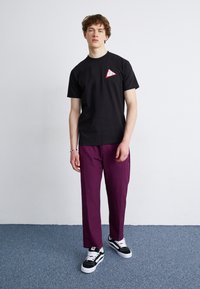 Camiseta de algodón negra con un logo en forma de triángulo en rojo y blanco, combinada con pantalones morados de corte holgado y zapatillas negras y blancas.