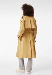 Manteau trench en khaki avec un design ajusté, taille cintrée et épaulettes. Texture lisse, longueur jusqu'aux genoux, accents de boutons sur les manches.