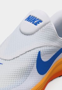 Λευκό υφασμάτινο αθλητικό παπούτσι Nike με μπλε λογότυπο Swoosh και μπλε κείμενο "NIKE" στη γλώσσα, με πορτοκαλί και κίτρινη σόλα.