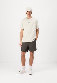 Camiseta de color beige claro con mangas cortas y un pequeño logo impreso, combinada con pantalones cortos gris oscuro y zapatillas blancas, rematada con una gorra clara.