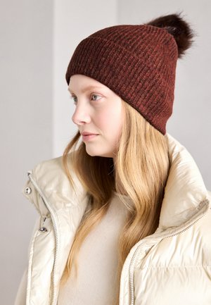 Cappello lavorato a maglia color bordeaux con texture a costine e un pompon in pelliccia sintetica sulla sommità. Indossato con un giubbotto imbottito avorio e una maglietta color crema.