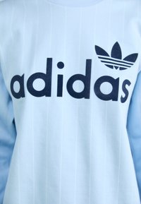 Sudadera azul claro con rayas verticales, presenta un gran logo negro de "adidas" y el icónico emblema de las tres rayas en el pecho.