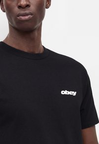 Άνδρας που φοράει μαύρο T-shirt με λευκό λογότυπο "obey" στο αριστερό στήθος, μπροστά σε απλό φόντο.