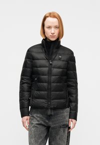 Jeune personne aux cheveux courts roux portant une doudoune noire et un jean noir délavé avec une déchirure sur la cuisse droite, debout devant un fond blanc.