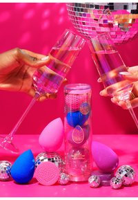 Set van kleurrijke make-upsponzen in roze, blauw en paarse kleuren, verpakt in een transparante cilinder met een sterrenpatroon, op een felroze achtergrond.