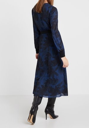 Femme portant une longue robe fleurie bleu foncé avec des poignets noirs et des bottes noires à talons hauts montant jusqu'aux genoux, debout de dos.