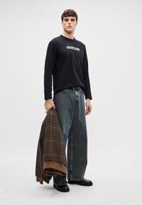 Camisola preta de manga longa com o texto "Calvin Klein", combinada com jeans azuis largos e uma jaqueta xadrez pendurada em um braço. Botas pretas.